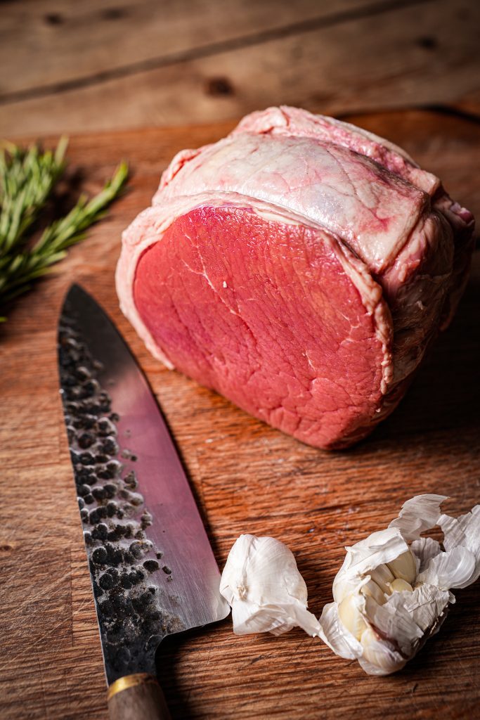 A raw rolled beef joint on a wooden board with a knife, rosemary and garlic